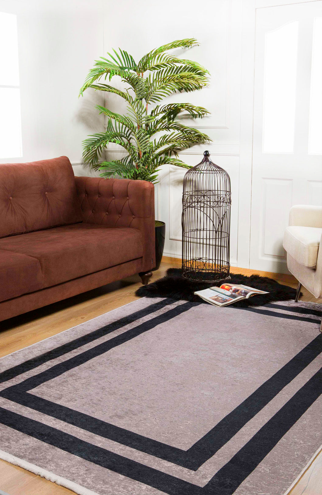 A cozy living room features a brown sofa, beige armchair, large green plant, empty black birdcage, and the Regal Trellis Washable Rug (JR601) in gray with black borders. A magazine lies nearby. White walls and wood floors complete this allergy-sensitive space.