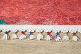 Close-up of the Rosy Blush - Machine-Washable Rug (JRS5067): red rug edge with cream tassels, some tied with red, blue, or yellow thread. Its vibrant red body features a hint of green in one corner and rests on a wooden floor.