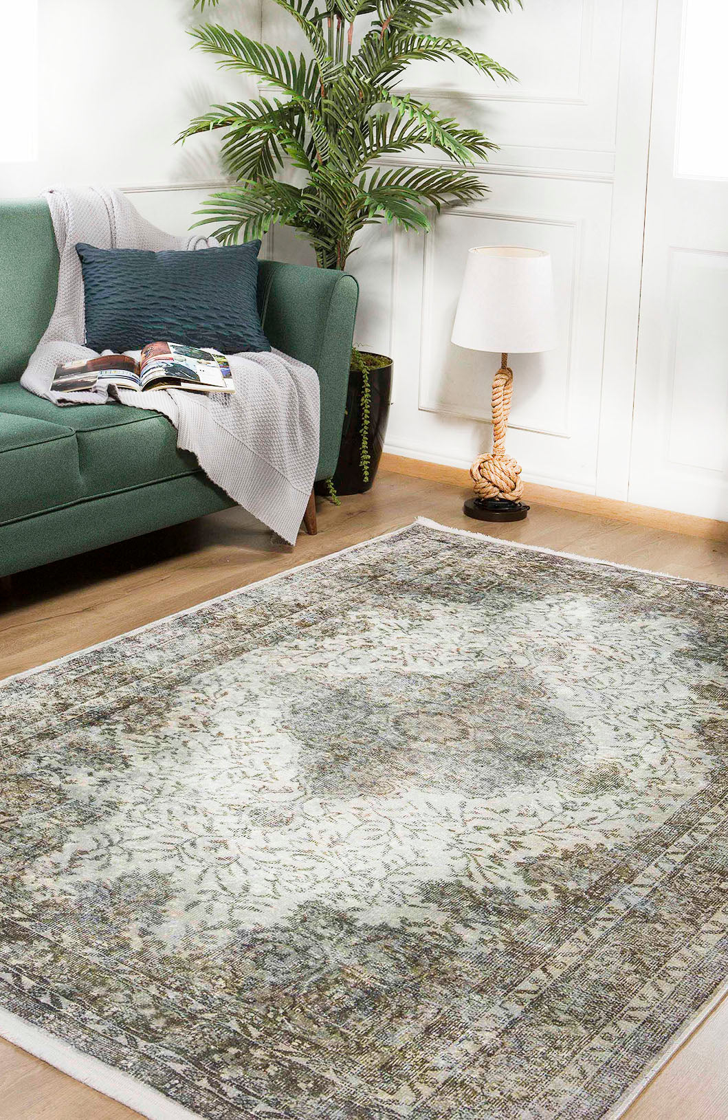 A living room with a green sofa, gray throw, and open magazine sits by a leafy plant and rope-wrapped lamp. The wooden floor is covered by the Royal Legacy - Washable Rug - JR207, ideal for high-traffic spaces.