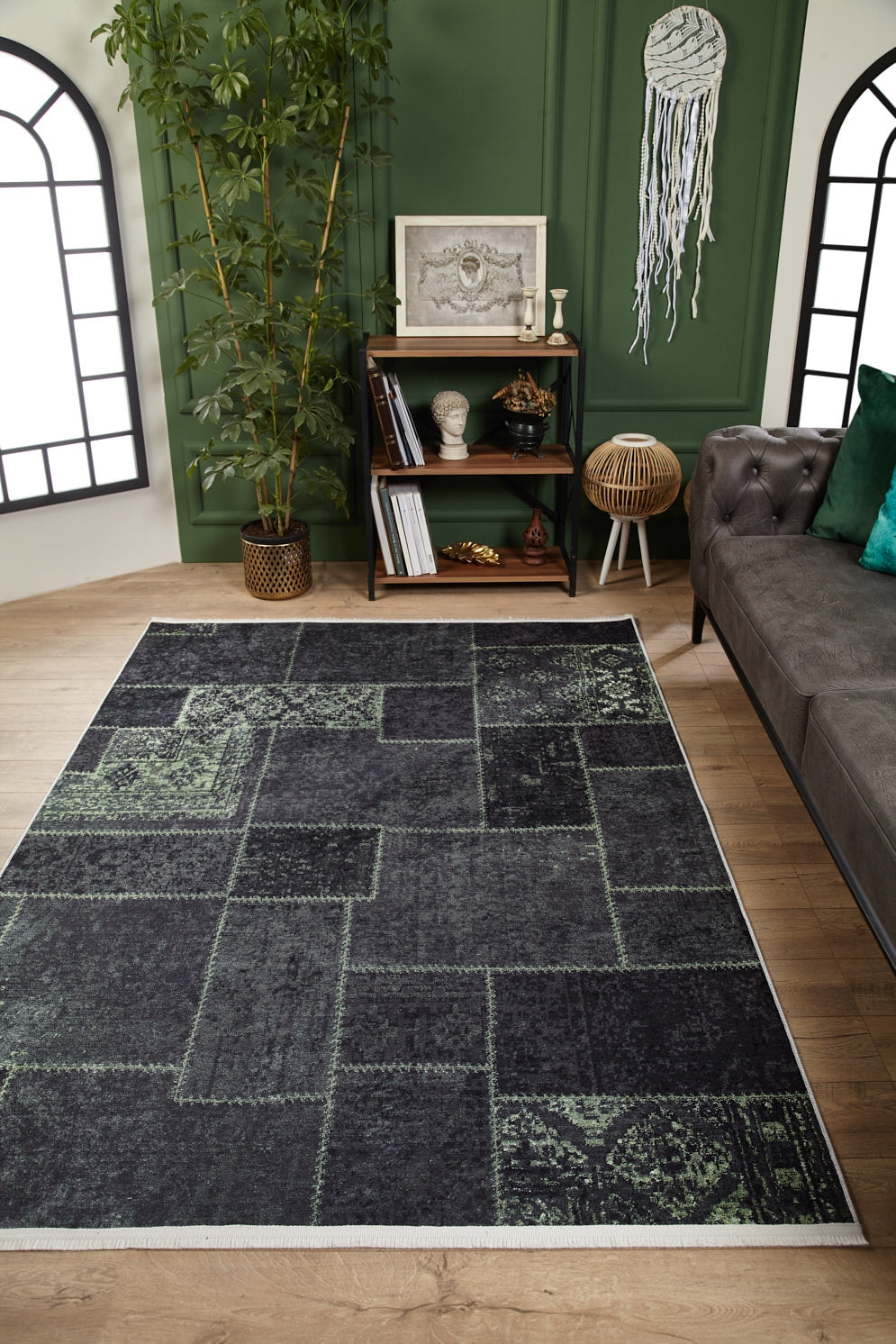 A modern living room with the Rustic Remix - Washable Rug - JR1710 in gray patchwork, perfect for high-traffic areas, wooden floors, a green accent wall, dark sofa, wooden decor shelf, potted bamboo plant, and geometric windows with natural light.