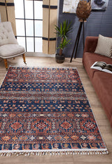 A living room featuring the Sahara Sunrise - Washable Rug - JR5091, ideal for high-traffic areas, a brown couch with an open book, a beige tufted chair, a potted plant, and large windows letting in natural light.