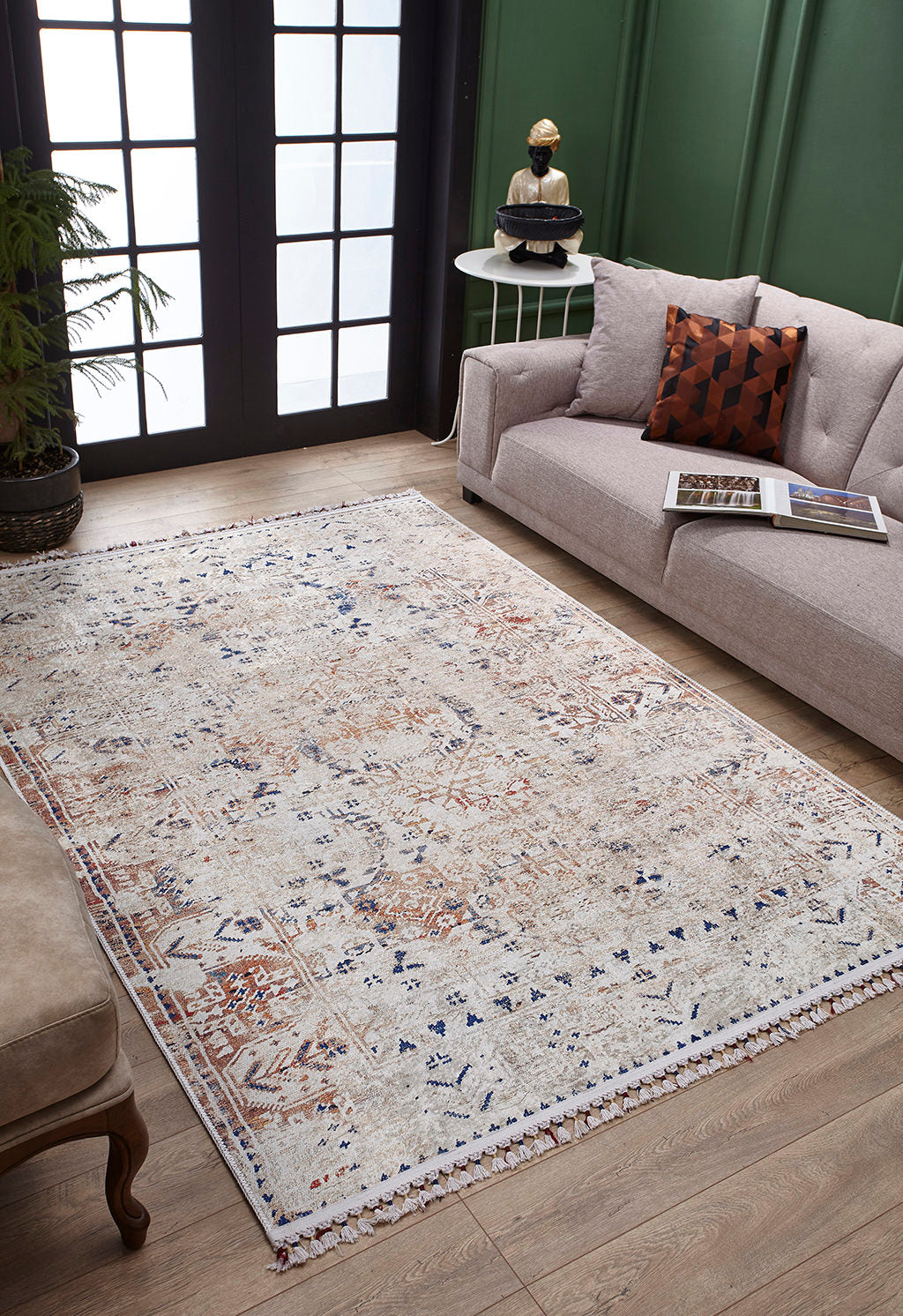 A cozy living room featuring the Shangri-La - Washable Rug - JR5042 in cream and beige, a beige couch with a geometric pillow, a round table, potted plant, and large black-framed windows—ideal for allergy-sensitive homes.