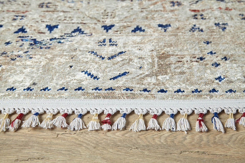 Load image 1 in gallery view. A close-up of the Shangri-La - Washable Rug - JR5042 in cream and beige with blue patterns and a red, blue, and white tasseled fringe, placed on a wooden floor—ideal for allergy-sensitive homes.