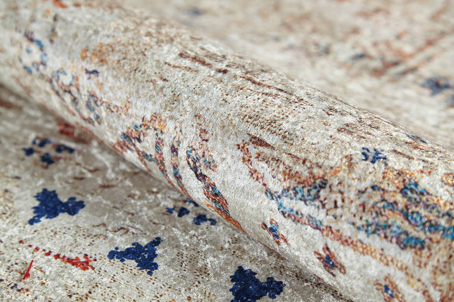A close-up of the Shangri-La - Washable Rug - JR5042 shows its rolled, allergy-sensitive cream and beige textured surface with subtle blue, red, and brown patterns. The soft, slightly shiny fabric makes it an ideal washable rug option.