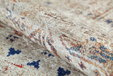 A close-up of the Shangri-La - Washable Rug - JR5042 shows its rolled, allergy-sensitive cream and beige textured surface with subtle blue, red, and brown patterns. The soft, slightly shiny fabric makes it an ideal washable rug option.