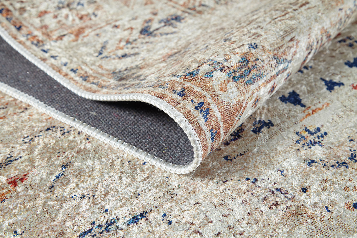 A close-up of the Shangri-La - Washable Rug - JR5042, featuring muted beige, blue, and rust tones with a textured surface; its edge is lifted to reveal a dark gray non-slip backing—perfect for allergy-sensitive spaces.