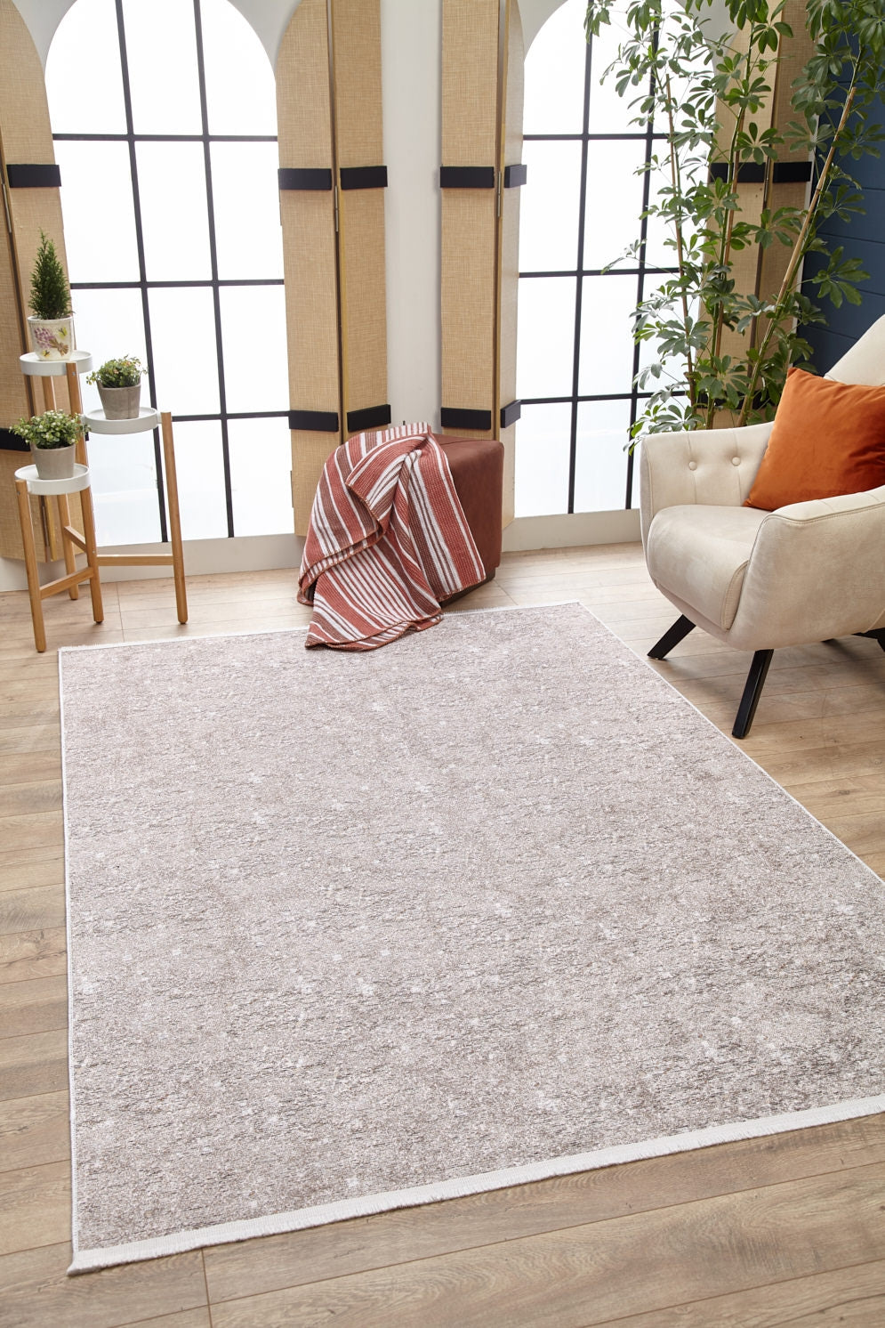 A modern living room with the Slate Charm - Washable Rug - JR1740 in light gray on wooden floors, a cream armchair with rust pillow, striped blanket on pouf, plant stand, and tall arched windows—ideal for high-traffic areas.