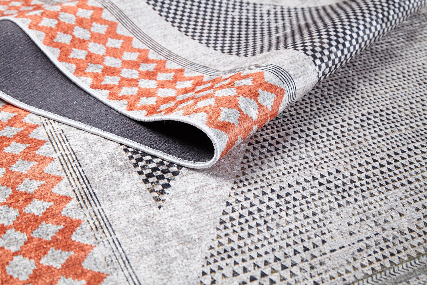 A close-up of the Sleek Design - Washable Rug - JR1727 shows orange and black geometric patterns on a light background with a folded corner revealing its texture—perfect for allergy-sensitive, high-traffic areas.