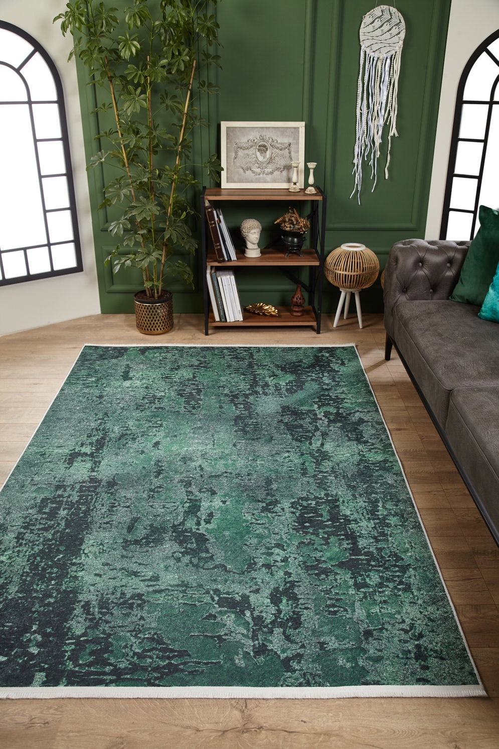 A modern living room featuring the Smokey Mountains - Washable Rug - JR1631 in green and black on wooden floors, paired with a gray tufted sofa, dark bookshelf, tall plant, green walls, large arched windows, and a wall hanging.