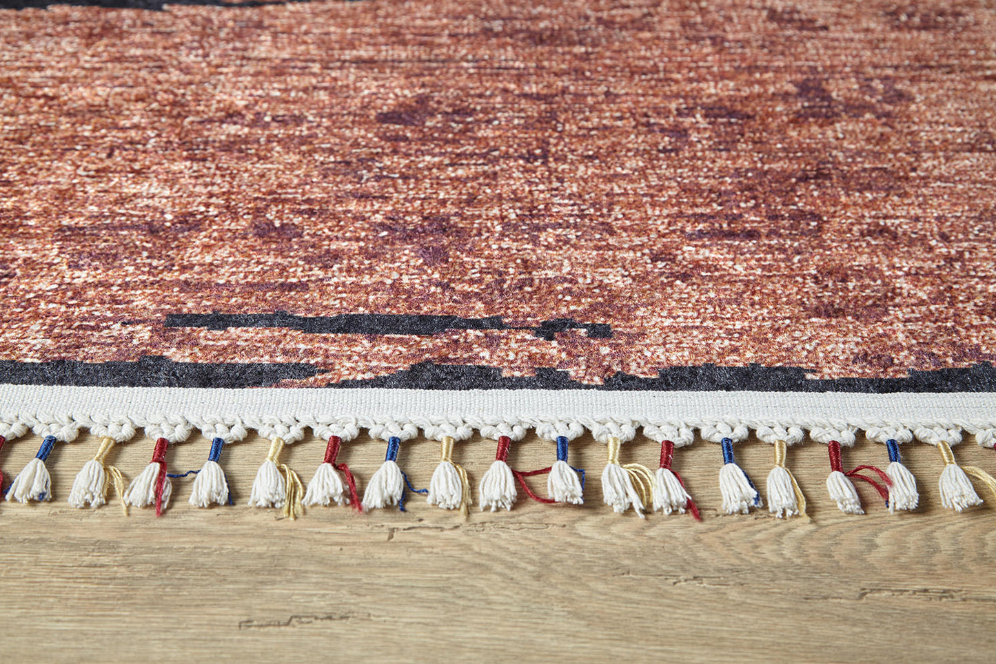 A close-up of the Soft Brushed - Washable Rug - JR5056 shows its textured reddish-brown pattern and white fringe with blue and red threads, making it a stylish, durable choice for high-traffic areas and wooden floors.