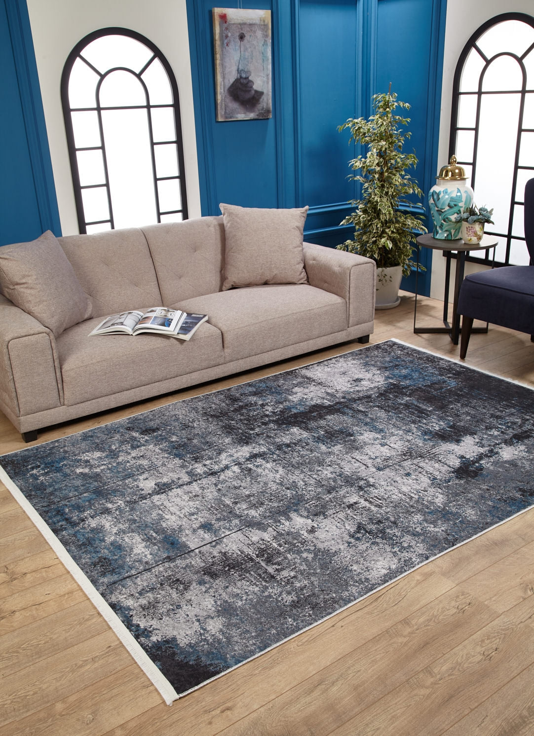 A beige sofa with cushions and an open book sits on a wooden floor beside the Solar Eclipse - Washable Rug - JR1921. Blue paneled walls, large windows, and modern decor create a bright, stylish living room ideal for high-traffic areas.