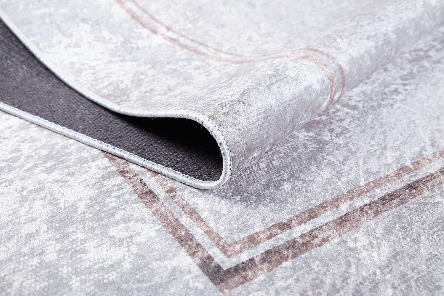 A close-up of the Sophisticated Edging - Washable Rug - JR1738 shows its light gray flat weave with a subtle brown border. One corner is folded to reveal the black non-slip backing—a practical, stylish choice for any space.