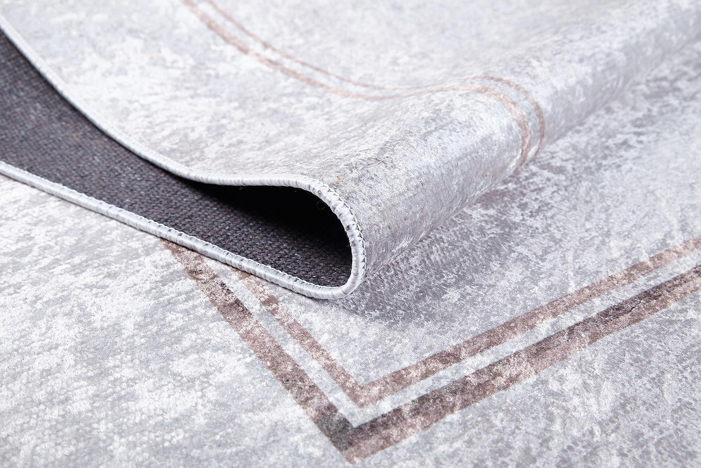 A close-up of the Sophisticated Edging - Washable Rug - JR1738 shows its light gray flat weave with a subtle brown border. One corner is folded to reveal the black non-slip backing—a practical, stylish choice for any space.