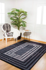 A modern living room showcases the Sophisticated and Chic - Washable Rug - JR1409, a geometric black flat-weave rug, paired with a white ornate chair, black-and-white cushion, fur throw, round side table, potted plant, and wood floors.