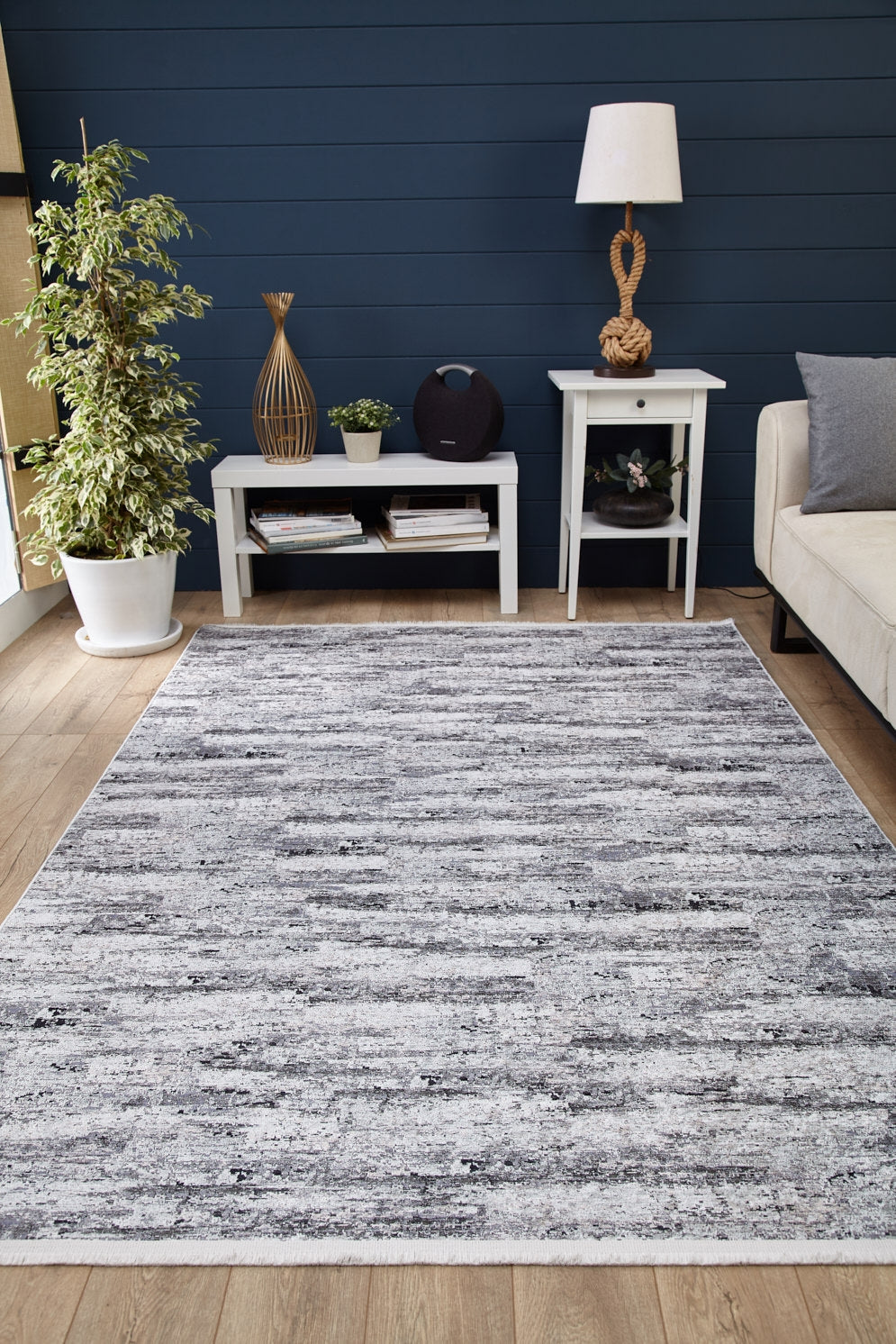 A modern living room with wood flooring features the Stone Path - Washable Rug - JR1772 (gray-anthracite, perfect for high-traffic areas), a white side table and lamp, small sofa, potted plant, and a blue shiplap accent wall.