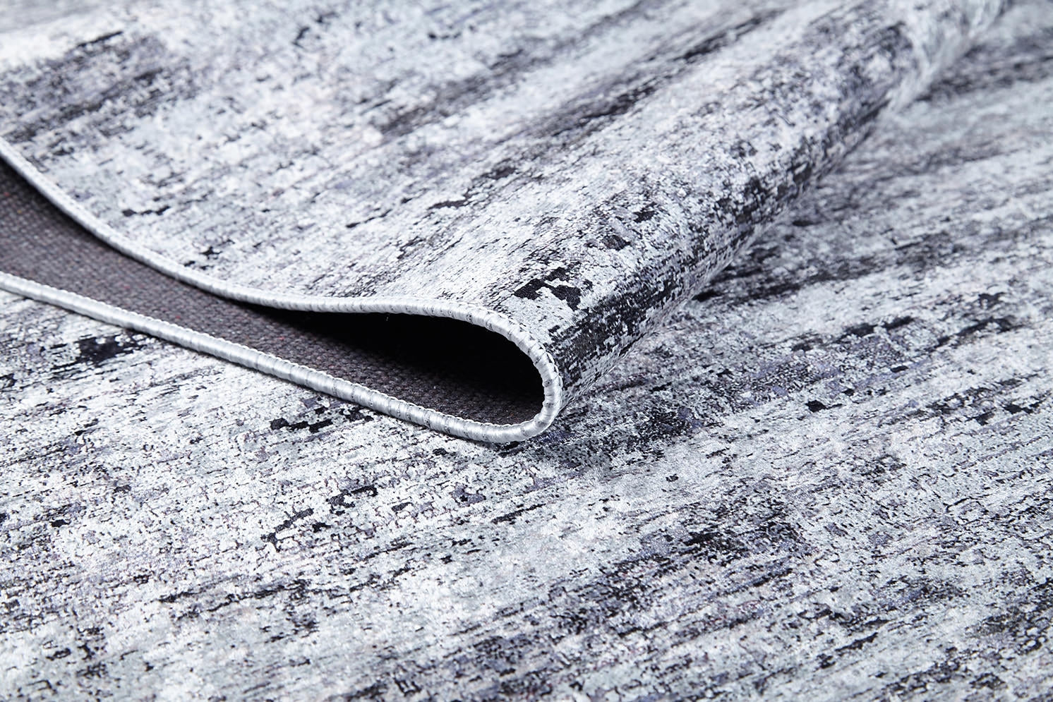 A close-up of the Stone Path - Washable Rug - JR1772, partially folded to reveal its soft surface and woven backing. Its distressed pattern in gray and black shades is perfect for high-traffic areas.