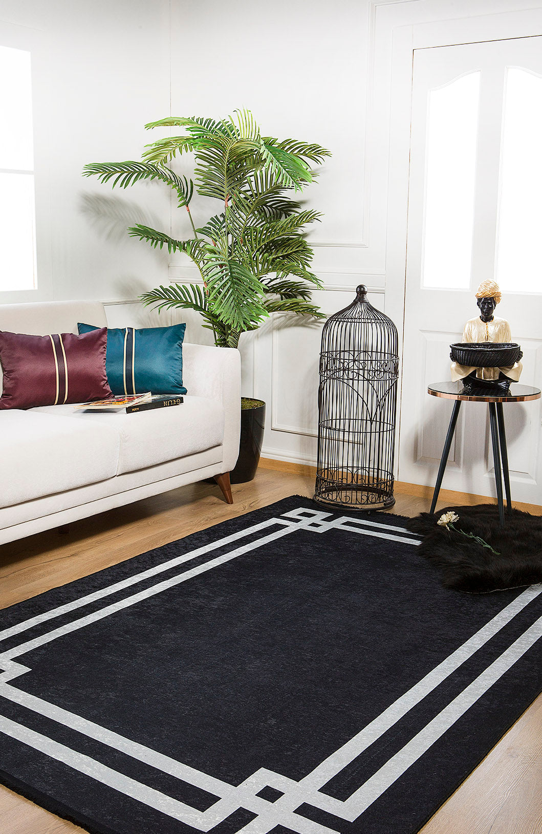 A modern living room featuring a white sofa with colorful cushions, a large green potted plant, the Subtle Border - Washable Rug - JR597 in black—perfect for high-traffic areas—a round side table, and an empty black birdcage.