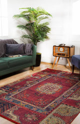 A cozy living room features a green sofa, vintage radio on a wooden stand, tall potted plant, and the Tapestry Tango - Washable Rug - JR313 in red—perfect for high-traffic areas—on a wooden floor with sunlight streaming from the left.