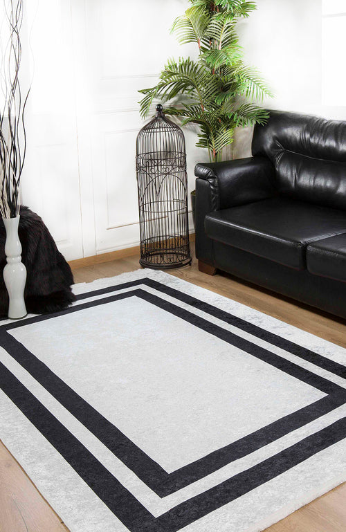 Load image 1 in gallery view. A modern living room with a black leather sofa, the Timeless Border - Washable Rug - JR572 in gray-anthracite, a tall green plant, an empty birdcage, a white vase with branches, and white walls with wooden floors.