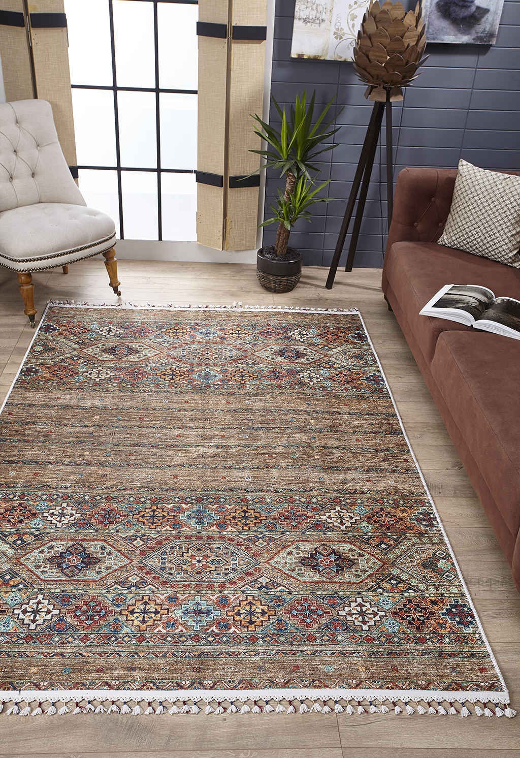 The Tribal Tango - Washable Rug - JR5089, featuring colorful geometric and floral patterns in bronze and brown, is laid on a wooden floor between a brown sofa, white tufted chair, and green plants in a modern living room.