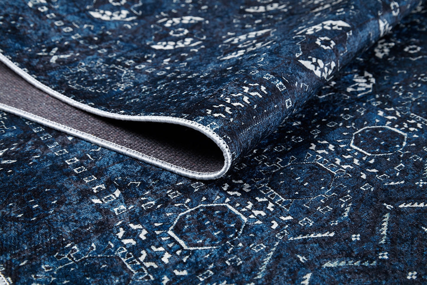 A close-up of the Tribal Trail - Washable Rug - JR1939 in blue and white, featuring intricate geometric patterns with a curled edge exposing the underside and texture—ideal for high-traffic spaces.