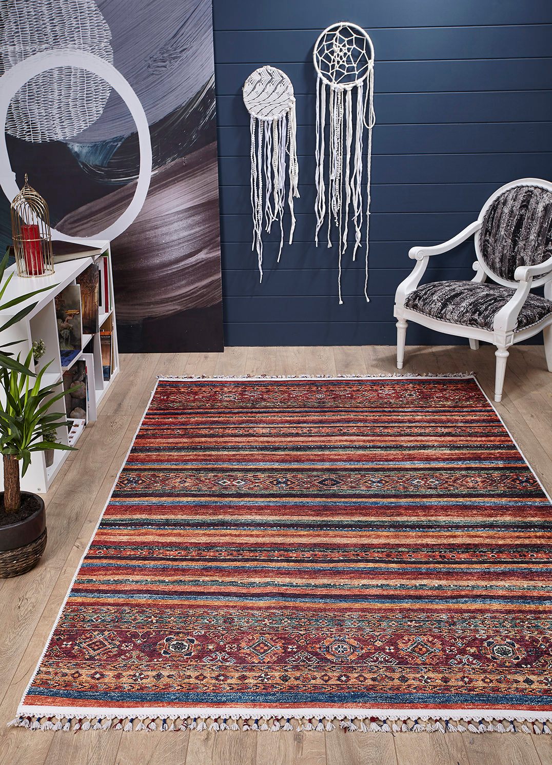 A room with the Tropic Thunder - Washable Rug - JR5077 (Outlet) US, ideal for high-traffic areas on a wood floor, a white armchair with a patterned cushion, dreamcatchers on a dark blue wall, a potted plant, and a white shelf with books and lantern.