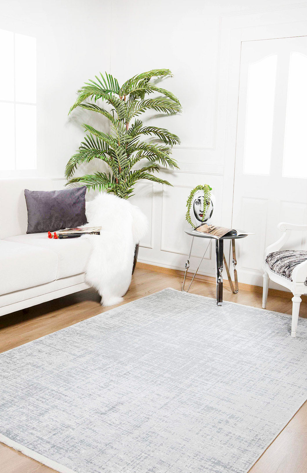 A bright, modern living room featuring a white sofa, gray pillow, faux fur throw, large potted plant, glass side table with books and a face sculpture, white armchair, and the Twilight Sky - Washable Rug - JR856 in gray-anthracite for high-traffic areas.