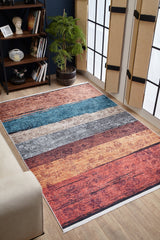 The Urban Edge - Washable Rug - JR2005, featuring multicolor horizontal stripes in blue, gray, orange, and red, adds a cozy touch to a living room with wooden floors, large bamboo-shaded windows, shelves, and a potted plant.