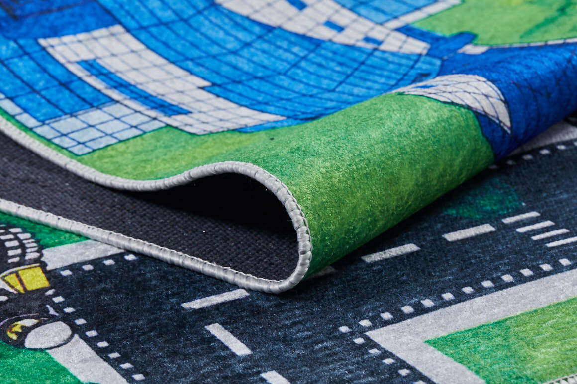 A close-up of the Urban Road - Machine-Washable Rug JRC013, a multicolor play mat with roads, grass, and buildings; one corner is curled up to show the flat weave texture and detailed edge stitching.