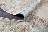 A close-up of the Urban Utopia - Washable Rug - JR1957 shows its folded corner, revealing a dark underside and stitched edges. This flat weave rug features muted beige, gray, and brown tones in a grid-like pattern.