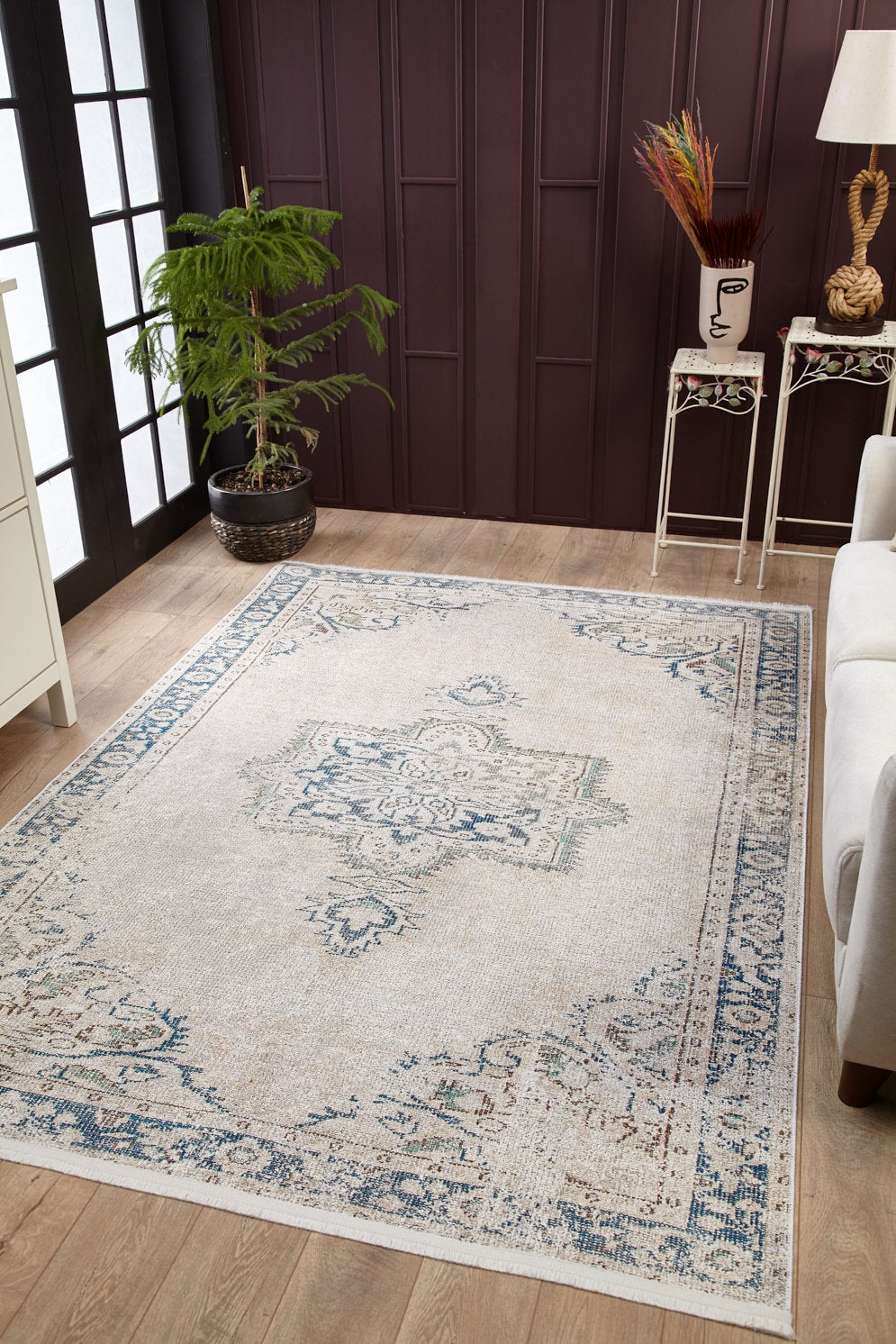 A living room featuring the Vintage Beauty - Washable Rug - JR1688 in beige and blue, paired with a white sofa, potted plant, side table with a vase and lamp, and dark wall paneling—ideal for allergy-prone or busy areas.