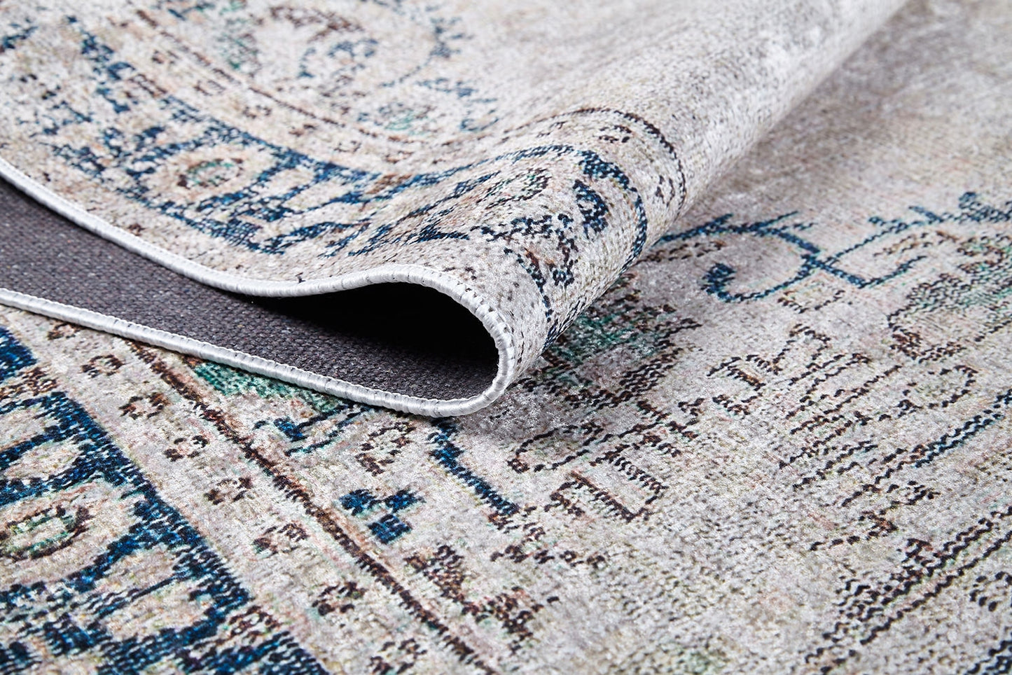 A close-up of the Vintage Beauty - Washable Rug - JR1688 with a lifted corner reveals its intricate blue, beige, and cream pattern and textured underside—ideal for high-traffic areas and allergy-sensitive homes.