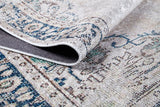 A close-up of the Vintage Beauty - Washable Rug - JR1688 with a lifted corner reveals its intricate blue, beige, and cream pattern and textured underside—ideal for high-traffic areas and allergy-sensitive homes.
