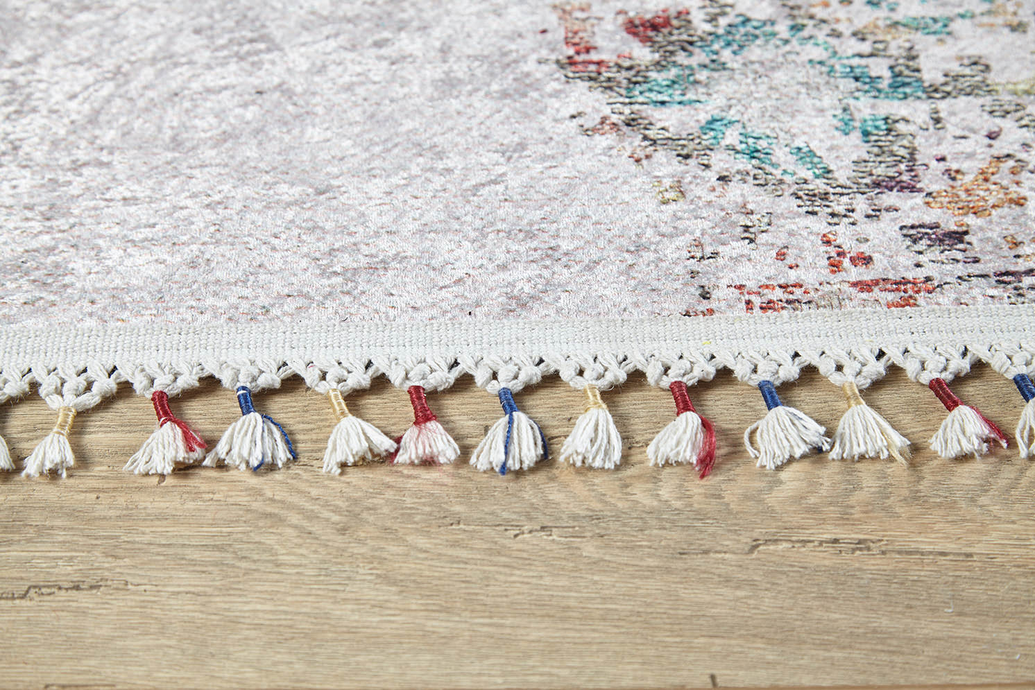 Close-up of the Vintage Cream Machine-Washable Rug (JR5006) in cream and beige, with multicolored tassels on the edge. This flat weave rug has a subtle pattern featuring hints of blue and red, shown resting on a wooden floor.