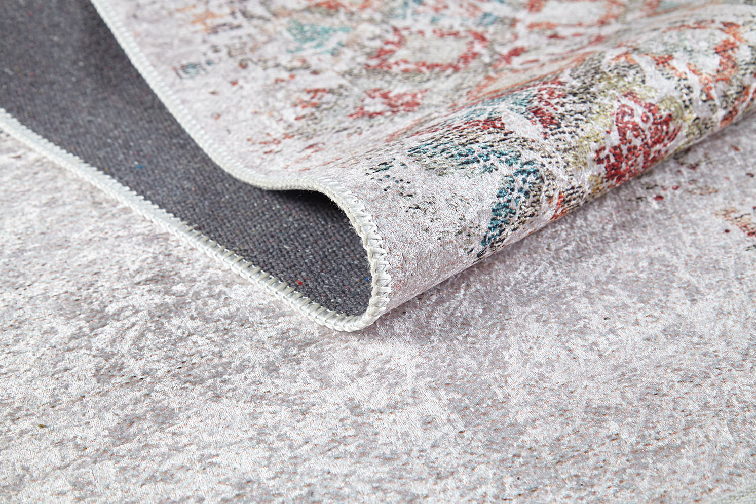 A close-up of the Vintage Cream - Machine-Washable Rug (JR5006) flipped over on a light gray textured floor, highlighting its woven underside and colorful topside design.