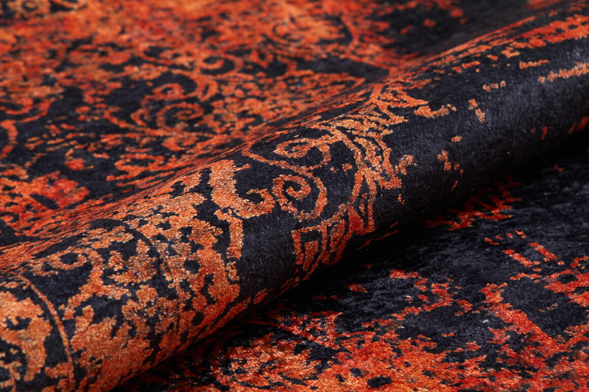 A close-up of the Vintage Glory - Washable Rug - JR1490 shows its ornate black and orange swirling and floral patterns on a textured surface. The partially rolled rug highlights its vibrant colors—ideal for high-traffic areas.