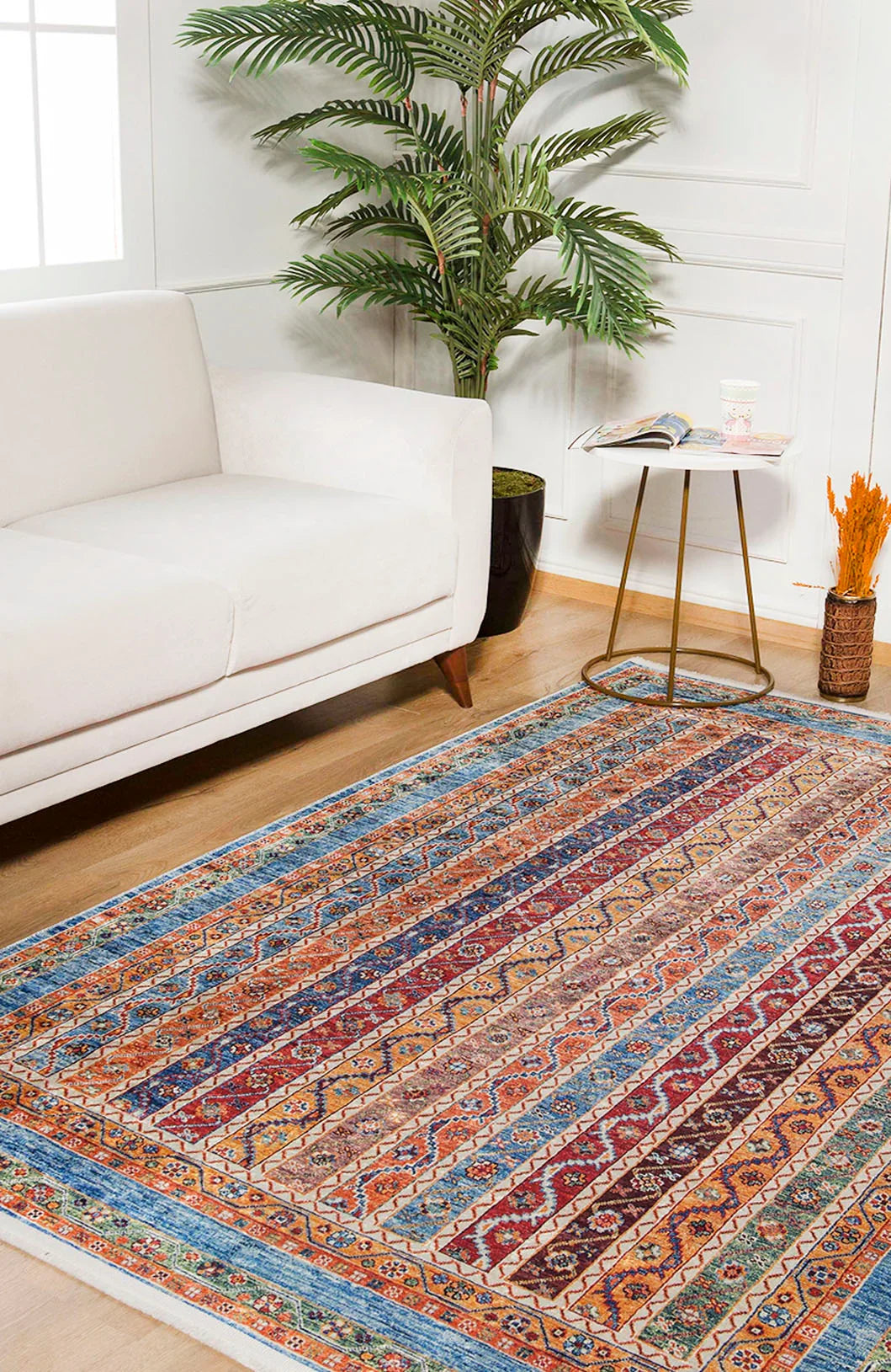 The Vintage Vibes - Washable Rug - JR37, featuring multicolor geometric designs, brightens the wood floor beside a white sofa, round gold side table with books, and a tall potted plant—ideal for high-traffic modern living rooms.