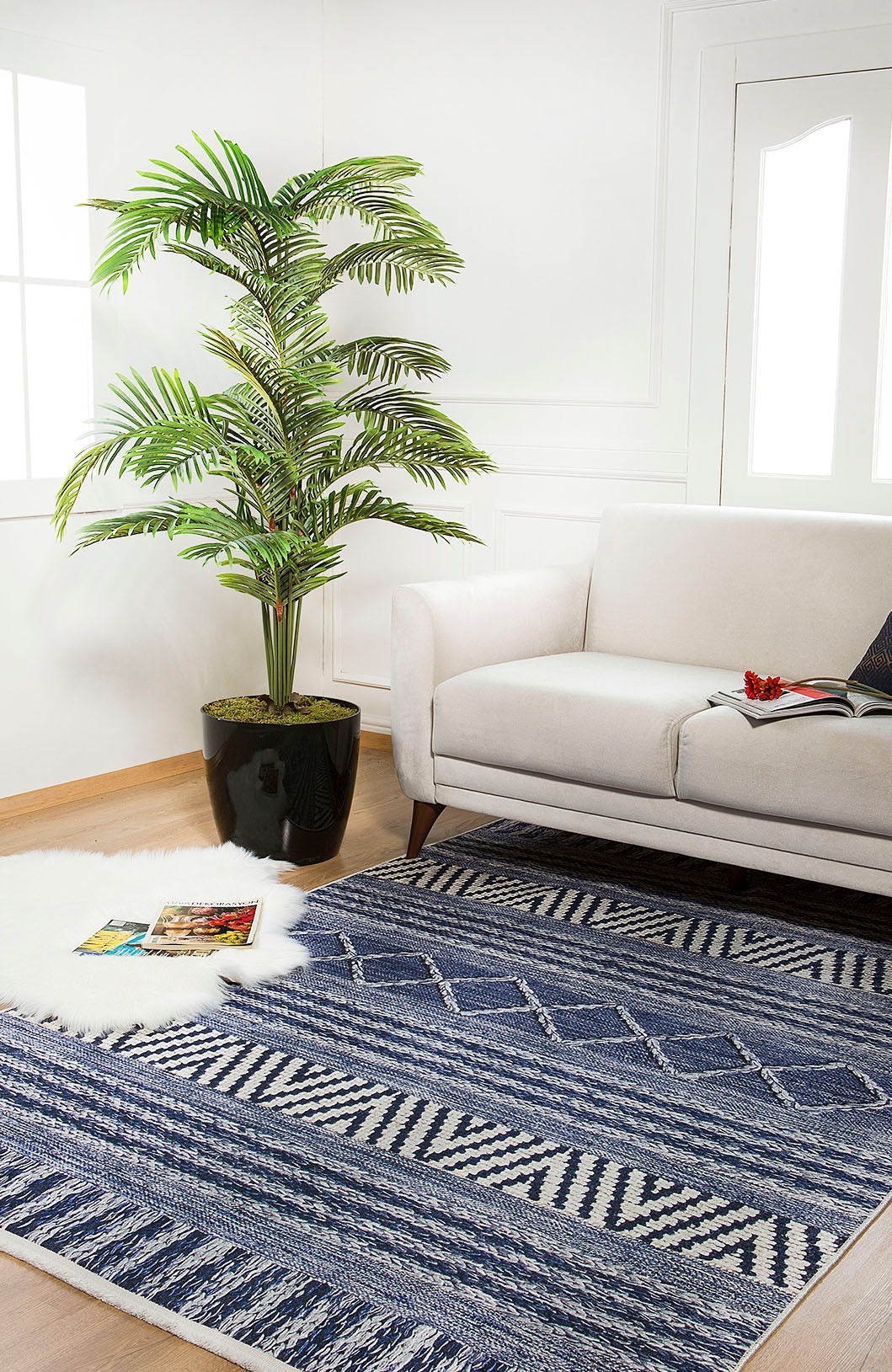 A bright living room features a white sofa, a large plant, and the Wavy Wonders - Washable Rug - JR941 in blue—perfect for high-traffic areas—with a fluffy white rug and magazines on light wooden floors by large windows.