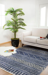 A bright living room features a white sofa, a large plant, and the Wavy Wonders - Washable Rug - JR941 in blue—perfect for high-traffic areas—with a fluffy white rug and magazines on light wooden floors by large windows.