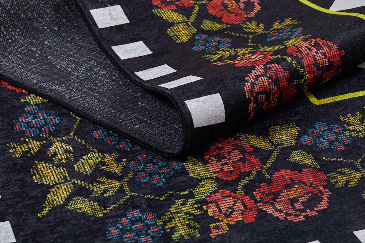 A close-up of the Wildflower Wonderland - Washable Rug - JR587 shows a black flat weave rug with red floral patterns, green leaves, white rectangular accents, a yellow border, and a slightly folded corner.