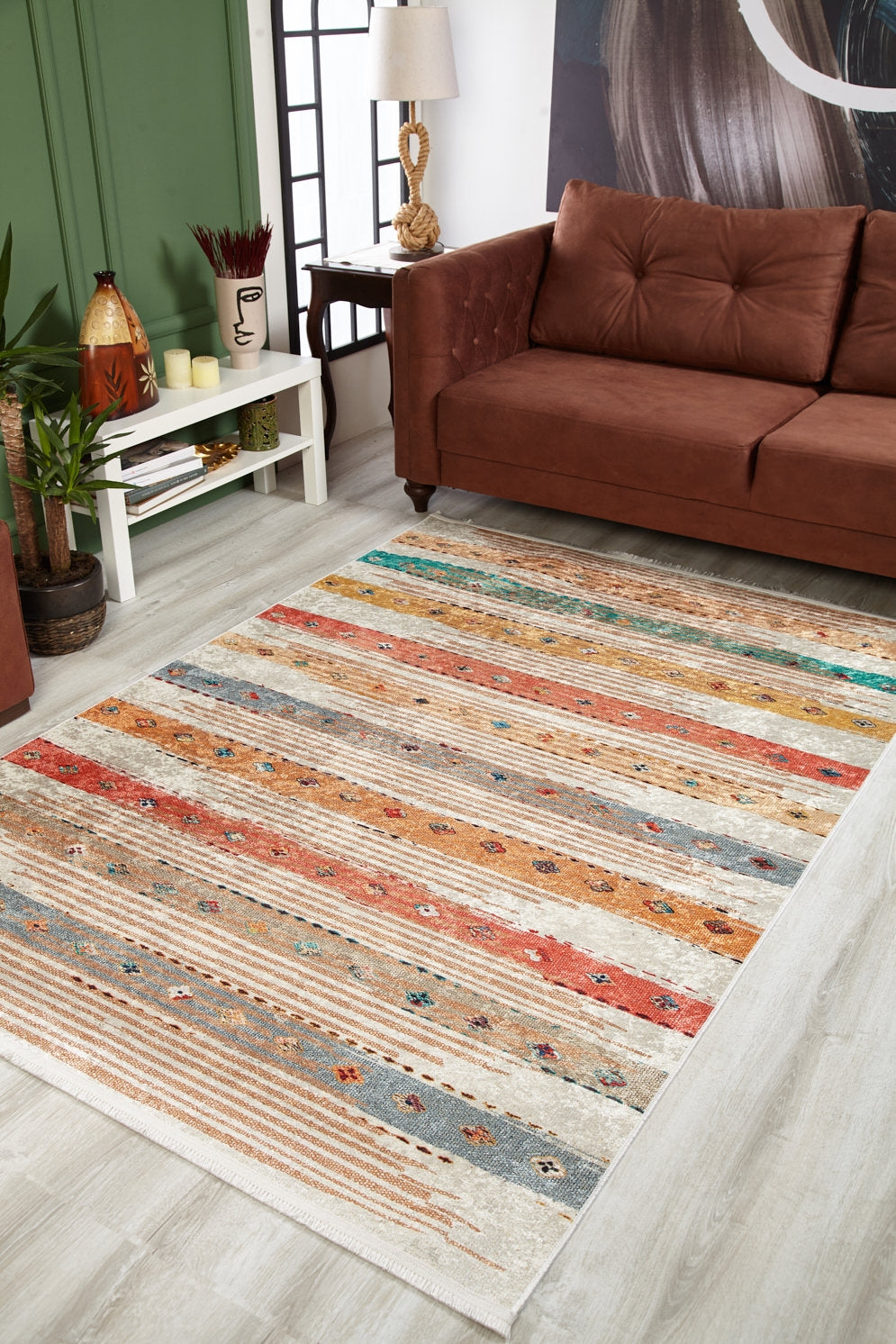 A living room with a brown sofa, green wall, side table with decor, and the Nomad's Nostalgia - Washable Rug - JR1669 showcasing multicolor subtle patterns on a light wood floor—ideal for high-traffic areas and easy care.
