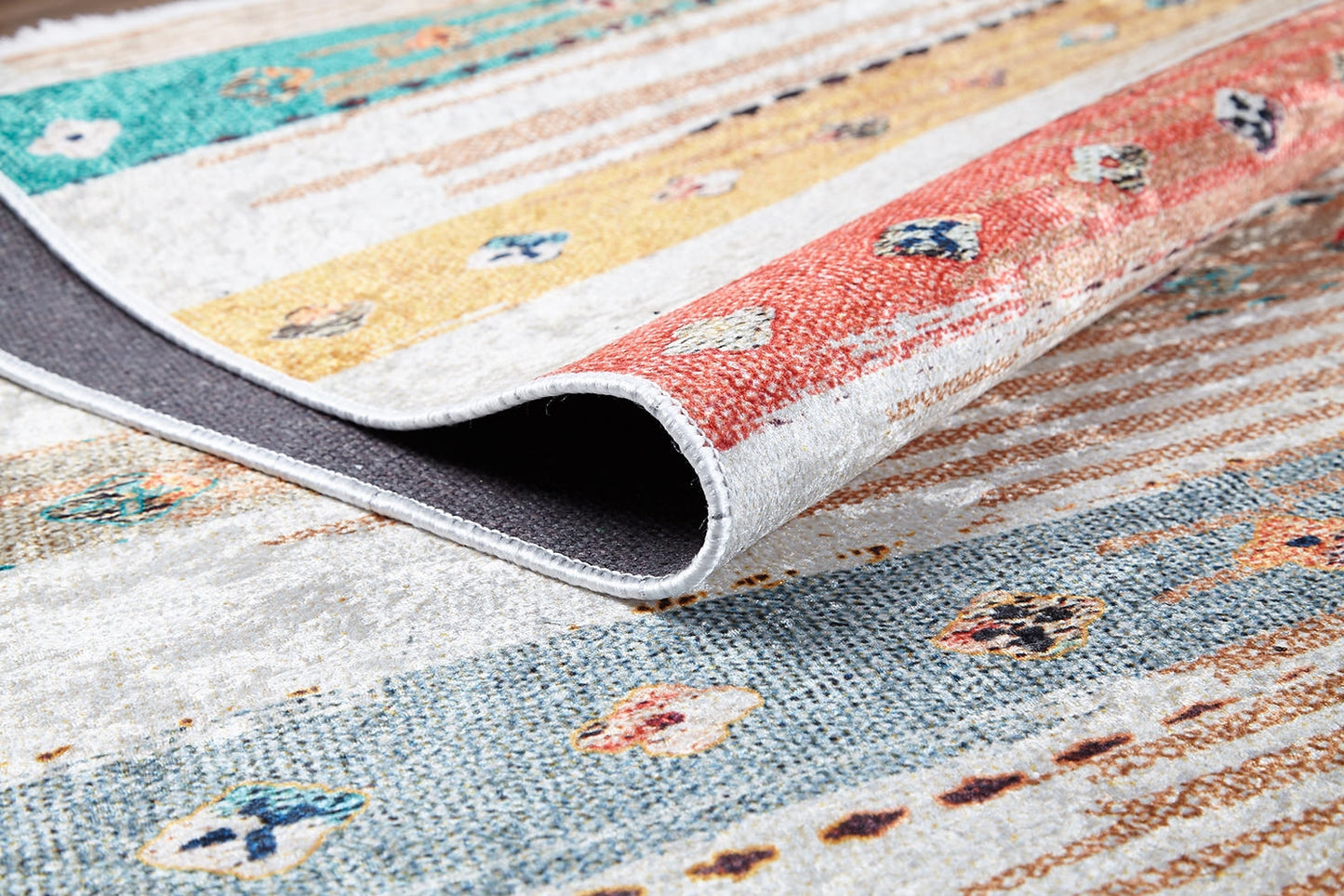 A close-up of the Nomad's Nostalgia - Washable Rug (JR1669) shows vibrant stripes and geometric shapes, with a folded corner revealing its dark underside—ideal for brightening high-traffic spaces.