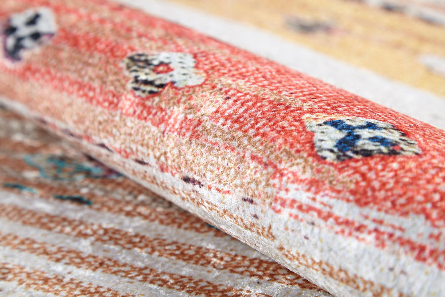 A close-up of the Nomad's Nostalgia - Washable Rug (JR1669) shows its rolled-up multicolor design featuring a red and beige pattern with blue and white floral accents, highlighting the rug's texture and intricate weave.
