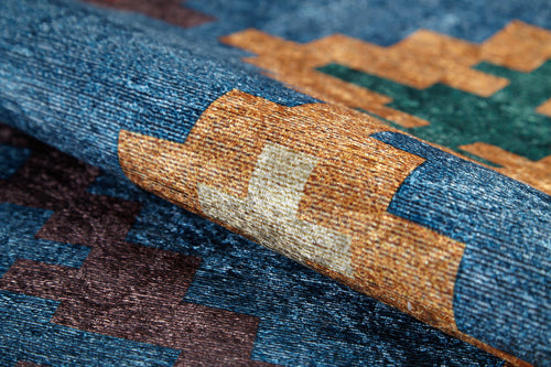 Load image 1 in gallery view. A close-up of the Nomad's Mirage - Washable Rug (JR5032), an allergy-friendly rug with a geometric pattern in blue, brown, gold, green, and cream, shown gently folded to highlight its textured design.