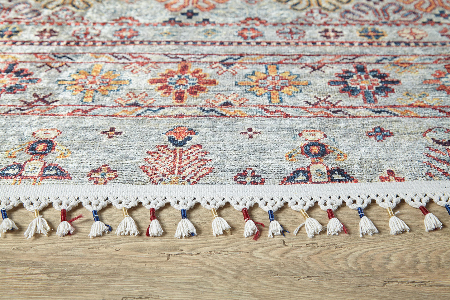 The Nomad's Nest - Washable Rug - JR5088 features floral and geometric patterns in red, blue, yellow, and orange with cream fringe tassels. Shown on a wooden floor, it's perfect for high-traffic areas and easy to clean.