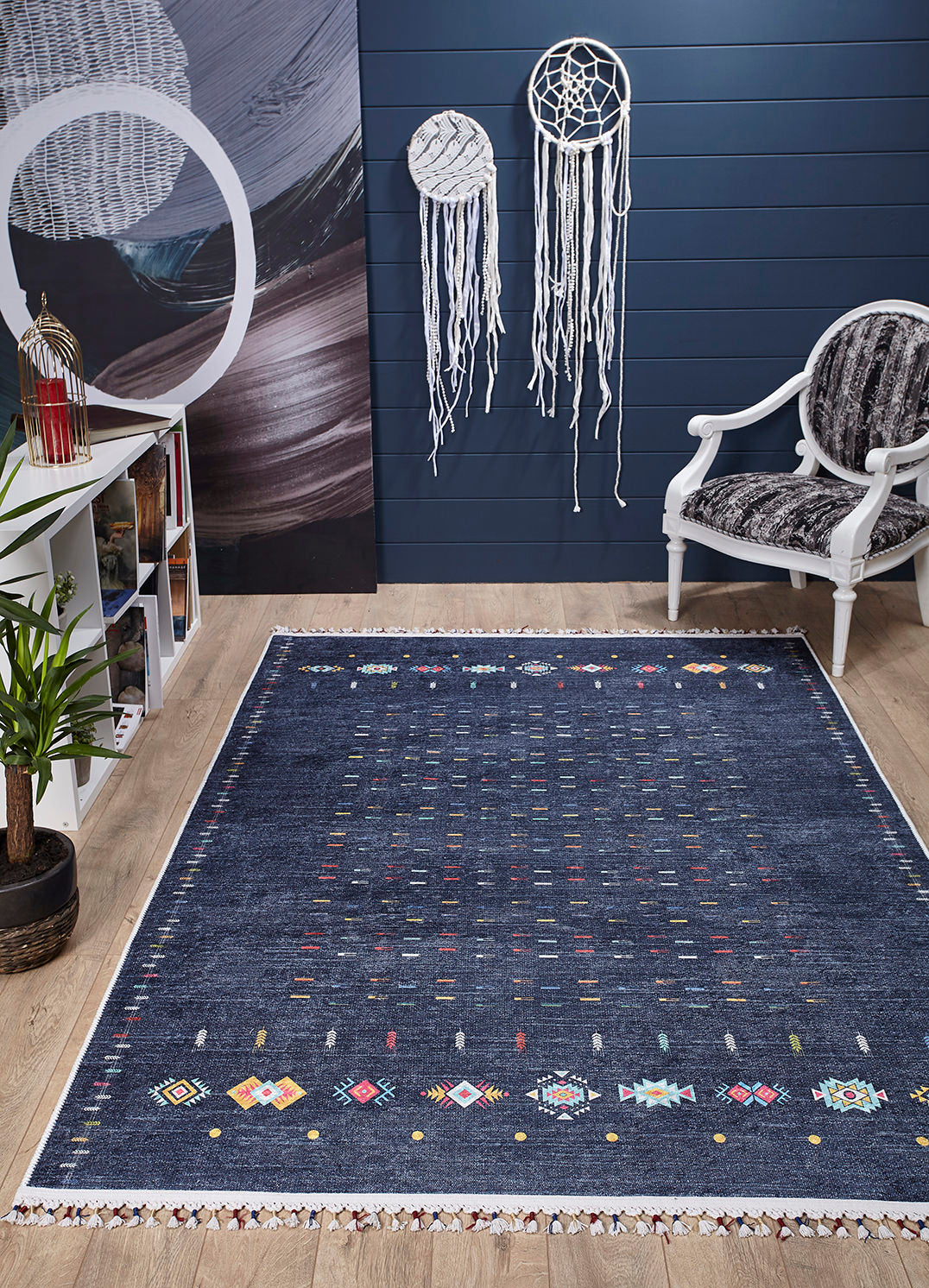 A cozy room features the Wanderer's Way - Washable Rug - JR5101 in dark blue, a white armchair, bookshelf, green potted plant, and two white dreamcatchers on a navy wall—perfect for high-traffic areas with its easy-care design. The floor is light wood.